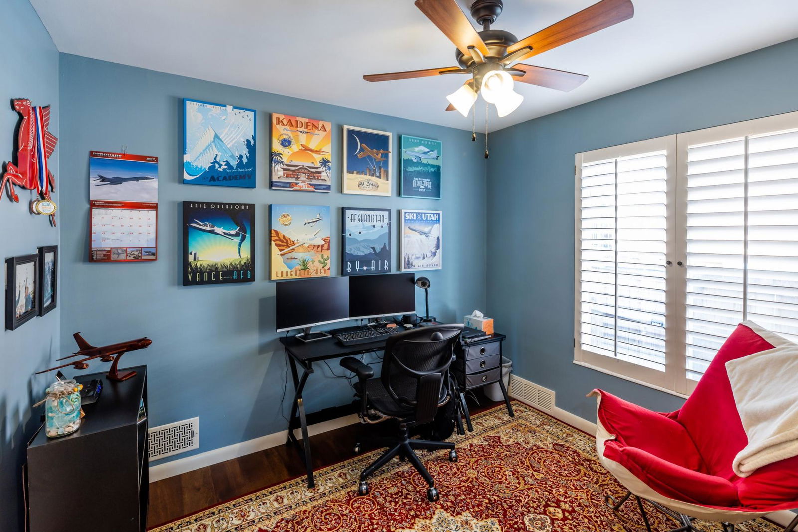 A room with a desk, chair, fan and pictures on the wall